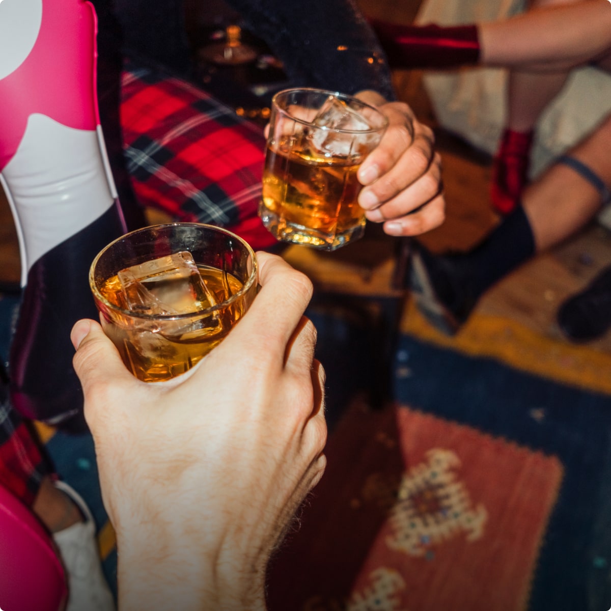 Two people clinking glasses of whiskey with ice cubes in a social setting.